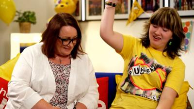 Two people seated on a sofa, with bright decorations and Pudsey Bear-themed items in the background.