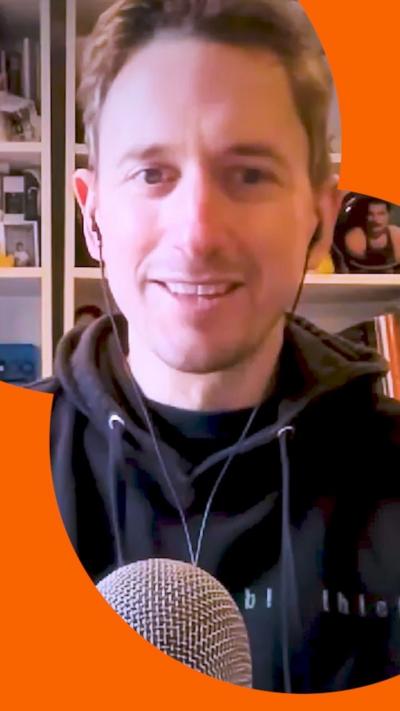 A man in a black hoodie sits behind a microphone in a room with a crowded shelf behind him