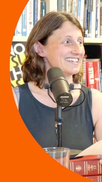 A woman with wavy brown hair sits behind a microphone. She is smiling at someone on her left. She is wearing a black tanktop.