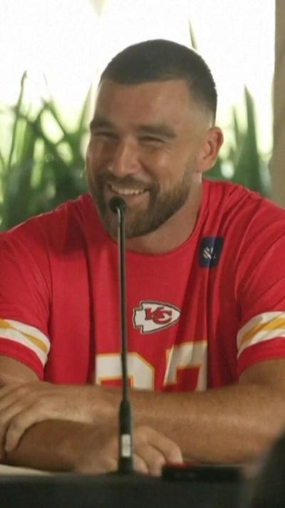 Travis Kelce behind a microphone wearing a Kansas City Chiefs shirt