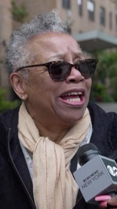 A woman with short grey hair and sunglasses speaks off camera, into a microphone being held near her.