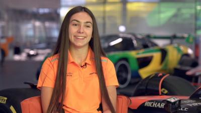 Ella Lloyd has long dark hair and is wearing a bright orange polo shirt with a McLaren logo