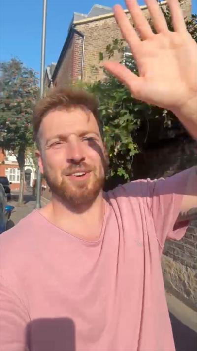 Man with blonde hair in pink t shirt waving at camera as he walks down residential street