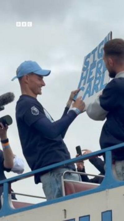 A man on top of an open top bus in a blue top and sky blue hat signs a card saying Sign Here Carl