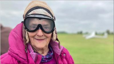 Barbara with flying goggles on smiling to camera with airfield in background.