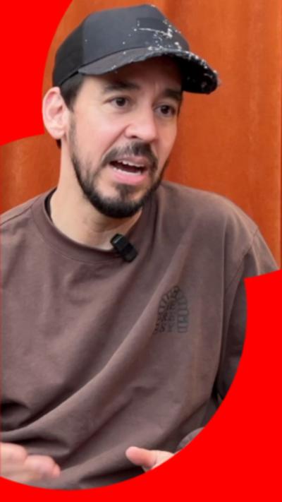 Mike Shinoda wearing a brown jumper and a black baseball cap. He's mid speech.