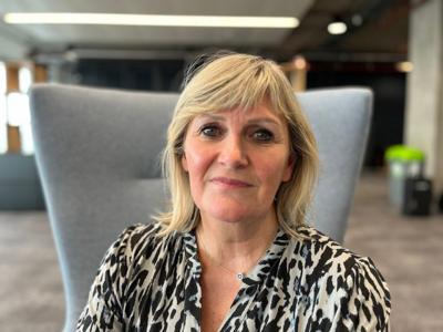 Angela MacVicar, with shoulder length blonde hair and wearing a patterned black and white blouse, sitting on a grey chair as she looks straight at the camera