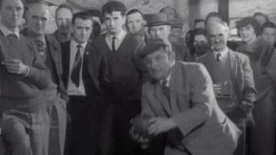 A man plays Scottish skittles, surrounded by a crowd of people