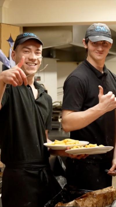 Two chip shop employees in black uniform and caps put thumbs up and smile.