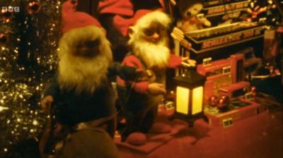 Model elves with white beards and Santa hats stand next to a Christmas tree in a display at a department store in 1978, One holds a glowing lamp. Toys including Scalextric sets can be seen.