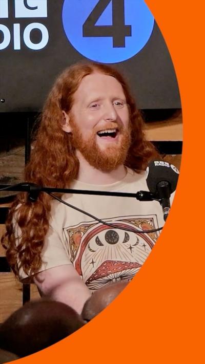 A man with long, flowing ginger hair sits behind a microphone. He is laughing. He is wearing a beige T-shirt with a mushroom design on it.
