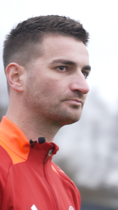 Man with short dark hair and red and orange sports top standing outside.