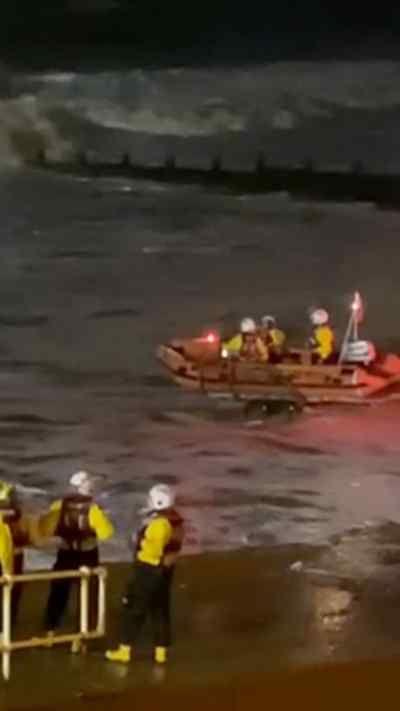 An orange rescue boat navigating through rough, choppy waters, with waves crashing around it. Several crew members wearing yellow jackets and white helmets are seated inside the boat.