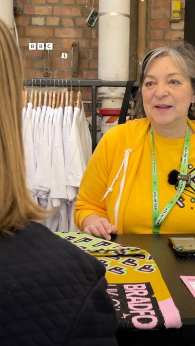 A woman in a yellow sweater looks across a counter at a woman, who is facing away from the camera.