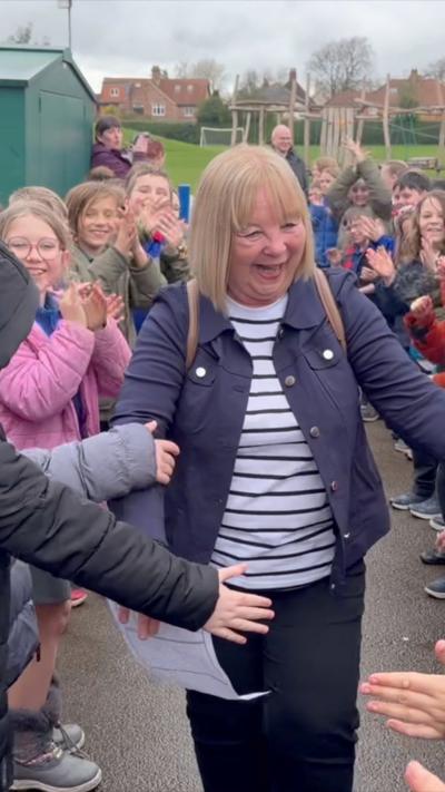 A woman in a blue jacket and stripy top walks through cheering children