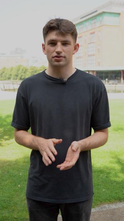 A young man with dark brown hair and wearing a black t-shirt. He is standing on a grassy area outside a university building. 