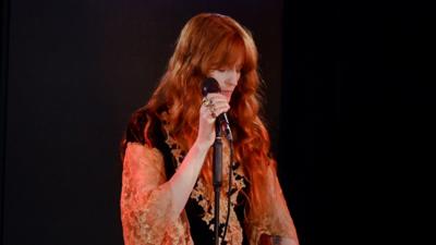Florence Welch stands behind a mic with her eyes closed. She wears a black and gold top 