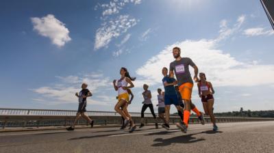 Blue sky and sunshine with light cloud above a groub of runners dressed in active wear and wearing race bibs 