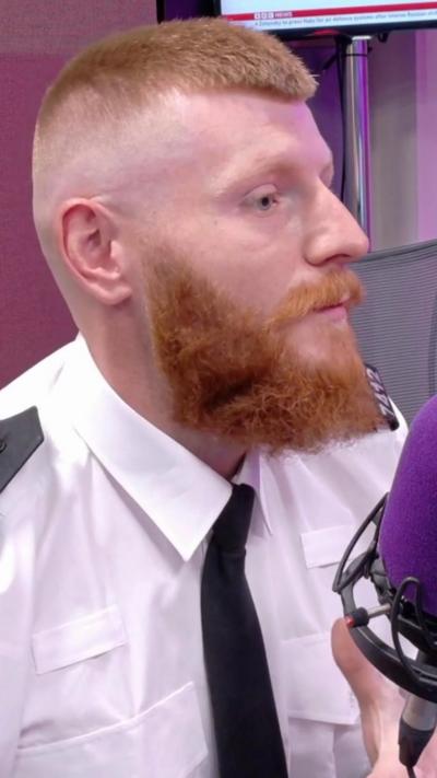 Charlie Jones has a ginger beard and short ginger hair. He wears a white and black police uniform. He looks at the camera, standing in front of a white wall.