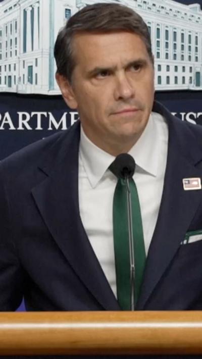 Todd Blanche, wearing a navy blue blazer and green tie, speaks from a podium and microphone with a logo of the Department of Justice behind him.