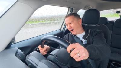 Weather presenter Matt Taylor, looking shocked with his mouth open as he grips the steering wheel of a small car, spinning around a skid pan.