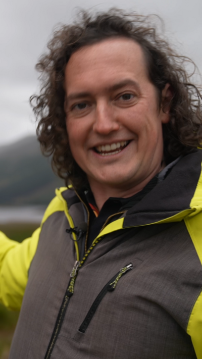 A man with long, curly hair and a black and yellow jacked stands in a valley with a loch in the Scottish Highlands.