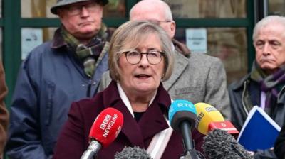 Andrea Nelson speaking into press mics, she has short blonde hair and is wearing a pair of black glasses. She has a burgundy coat on with a neutral scarf. Behind her are three men dressed in long coats and scarves.