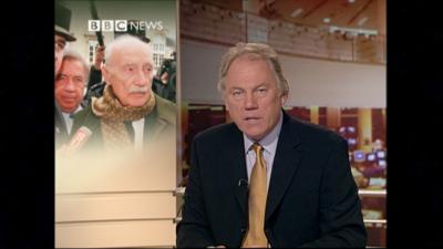 ý Newsreader Peter Sissons on the right of frame, facing the camera reading the news, wearing a dark yellow/mustard tie and a dark jacket.  The graphic behind on the left shows a picture of Maurice Papon surrounded by reporters and microphones, wearing a scarf.  There is a 'ý NEWS' graphic above the graphic.