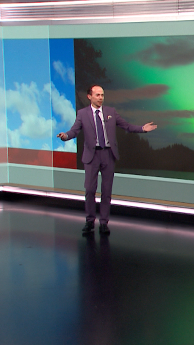 A weather presenter stands in front of images of the sky and northern lights. He has balding dark hair and is wearing a dark blue suit and tie.