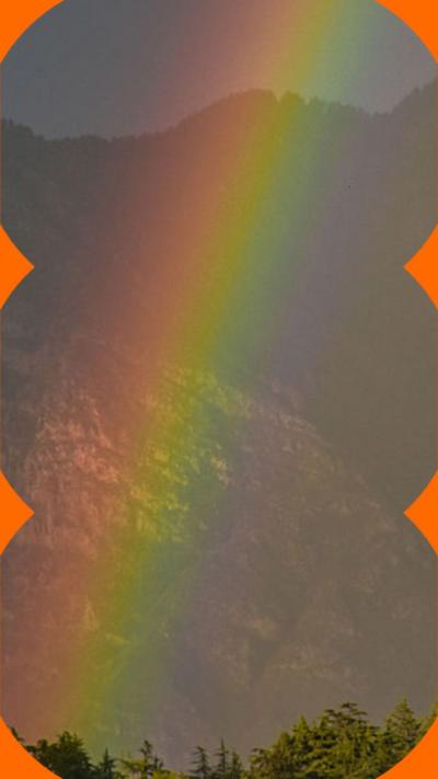 A rainbow is seen in front of a mountain. In the foreground is a hill with trees.