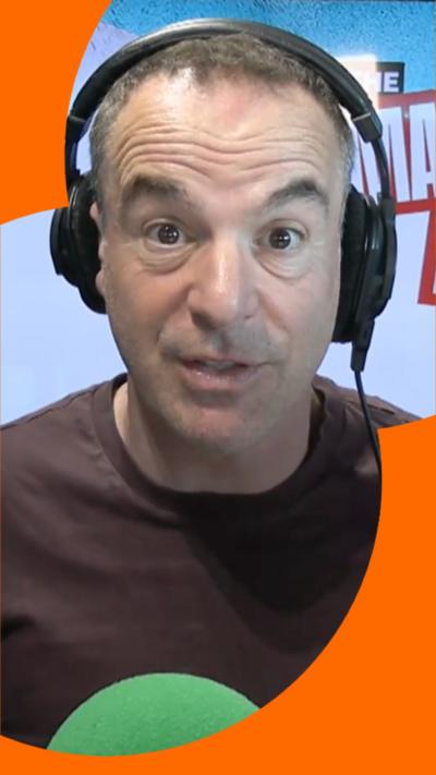 Martin Lewis wears headphones and a brown t-shirt. He sits in-front of a green microphone and looks directly at the camera.