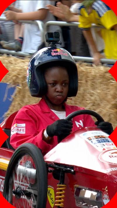 A young woman in red overalls and a crash helmet sits behind the steering wheel of a soapbox.