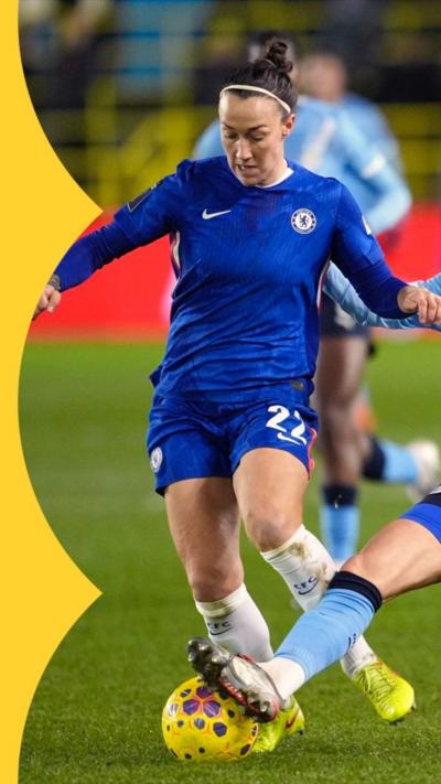 Two women, one in Chelsea footballer kit, one in Man City football kit, both try to kick a football