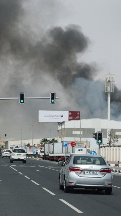 Cars drive down a road in Qatar towards a large billowing cloud of grey smoke coming from a missile strike