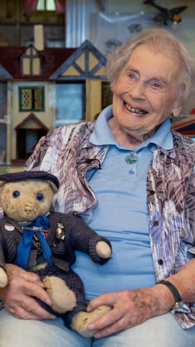 An elderly woman smiles while holding a vintage teddy bear in front of a doll's house display. 