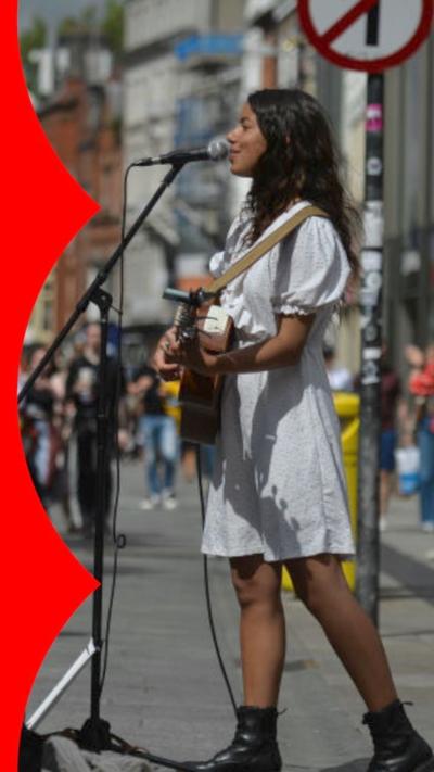 A woman in a white dress playing guitar and singing into a mic on the street.
