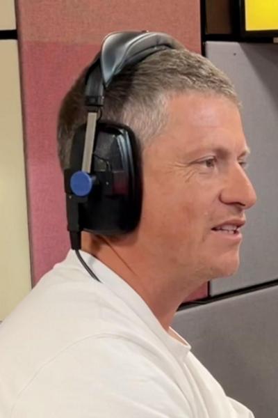Justin Rees, wearing a white T-shirt and wearing black headphones, is in a radio studio. He looks to the right, to someone off camera.