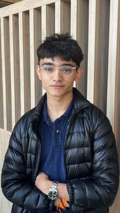 A teenager stands with his arms folded. He wears glasses and a dark jacket. He has short dark hair.