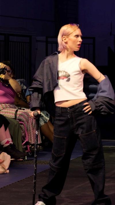 Person posing on a fashion runway in a white top and oversized dark trousers, holding a cane under stage lighting.