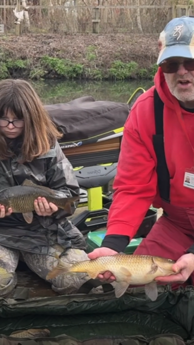 A man on the right wearing sunglasses, a grey cap and red hoodie, holding a fish out in front of him. On the left there is a girl with brown hair and glasses, wearing camouflage overalls and holding a smaller fish.