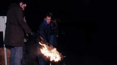 Two strikers dressed in warm coats and scarves stoke a fire on the picket line.