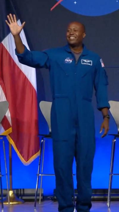 A man wearing a. blue jumpsuit standing on a stage waves to the crowd while smiling.