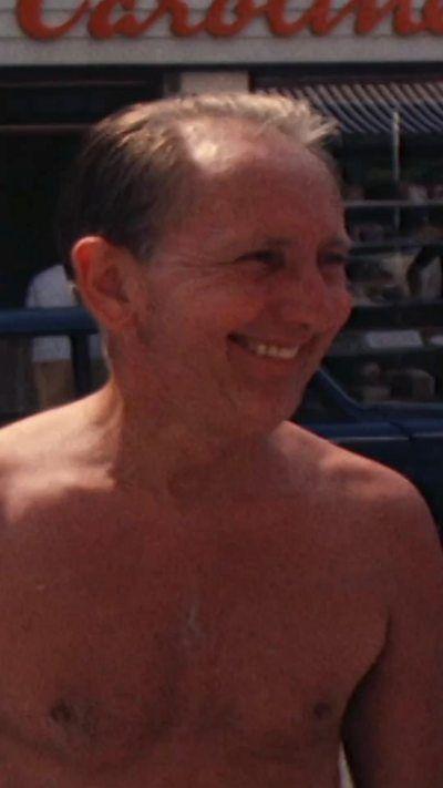 A man that has his top off in the sun. he is smiling at someone off camera. He has short dark hair.