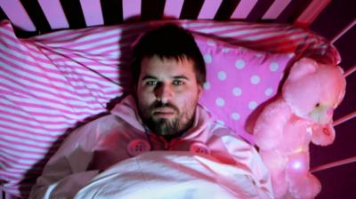 Man lies awake in bed surrounded by pink teddy and pink bed sheets