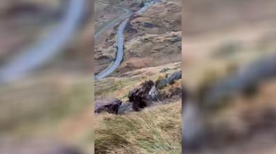 A boulder on a grassy hill with the A83 trunk road in the background.