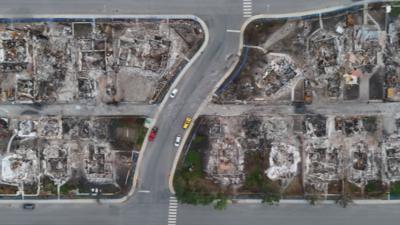 Several cars are parked on a street in between burned down houses.