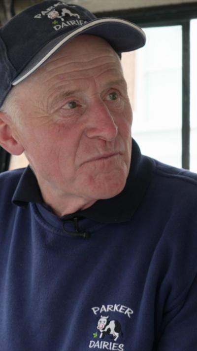 Close-up of an elderly man wearing a navy blue Parker Dairies cap and sweatshirt, looking slightly to the side.