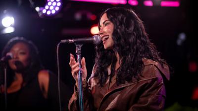 Demi Lovato sings into a mic in the Radio 1 Live Lounge. She has wavy dark hair and wears a brown jacket