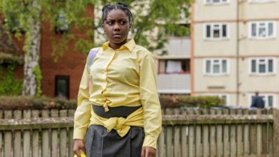 Girl with braided hair who is wearing a yellow shirt and grey skirt.