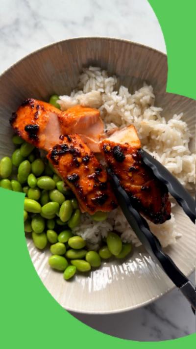 A bowl of rice, edamame beans and cooked salmon seen from above. A pair of tongs is holding a piece of the salmon.